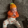 Jenson is registered to the contest to win money with this photo: baby, infant, orange_hat, striped_shirt, pumpkin_pants, couch, smile, cute, child, sitting, feet, clothing, face, headwear, patterned_pants, furniture, indoors, expression, playful, young_child