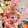 baby, child, infant, portrait, smiling, sitting, pink_bow, floral_dress, pumpkin, patterned_clothing, indoors, chair, bookshelf, books, playroom, play_mat, colorful, cozy, face, hands