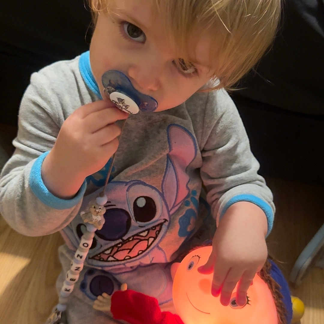 Kenzo participe au concours pour gagner de l'argent avec cette photo : bedroom, blonde_hair, child, cozy, cute, doll, floor, glowing_toy, indoor, pacifier, pajamas, person, playful, red_shoes, sitting, stitch, toddler, toy, wooden_floor, young_child