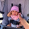 baby, child, stroller, pink_cap, pink_outfit, hand, finger, safety_strap, seat, happy, tongue_out, infant, cute, person, outdoor, sunlight, smile, clothing, baby_toy, holding
