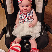 Renesmee is registered to the contest to win money with this photo: baby, child, stroller, headband, dress, smiling, indoor, footwear, white_skirt, red_shoes, cute, seated, infant, happy, person, colorful, patterned_clothing, safety_strap, car_seat, toddler