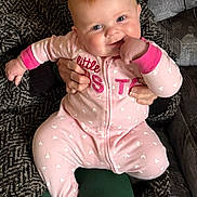 Carolina is registered to the contest to win money with this photo: baby, child, person, smile, pink_clothing, onesie, hands, couch, fabric, patterned_sweater, indoor, seated, happy, cute, face, skin, holding, fist, cozy, home
