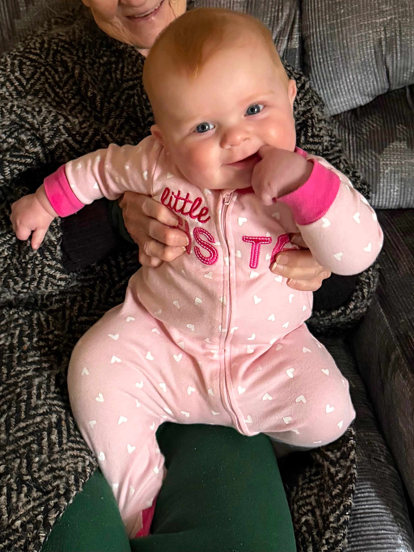 Carolina is registered to the contest to win money with this photo: baby, child, person, smile, pink_clothing, onesie, hands, couch, fabric, patterned_sweater, indoor, seated, happy, cute, face, skin, holding, fist, cozy, home