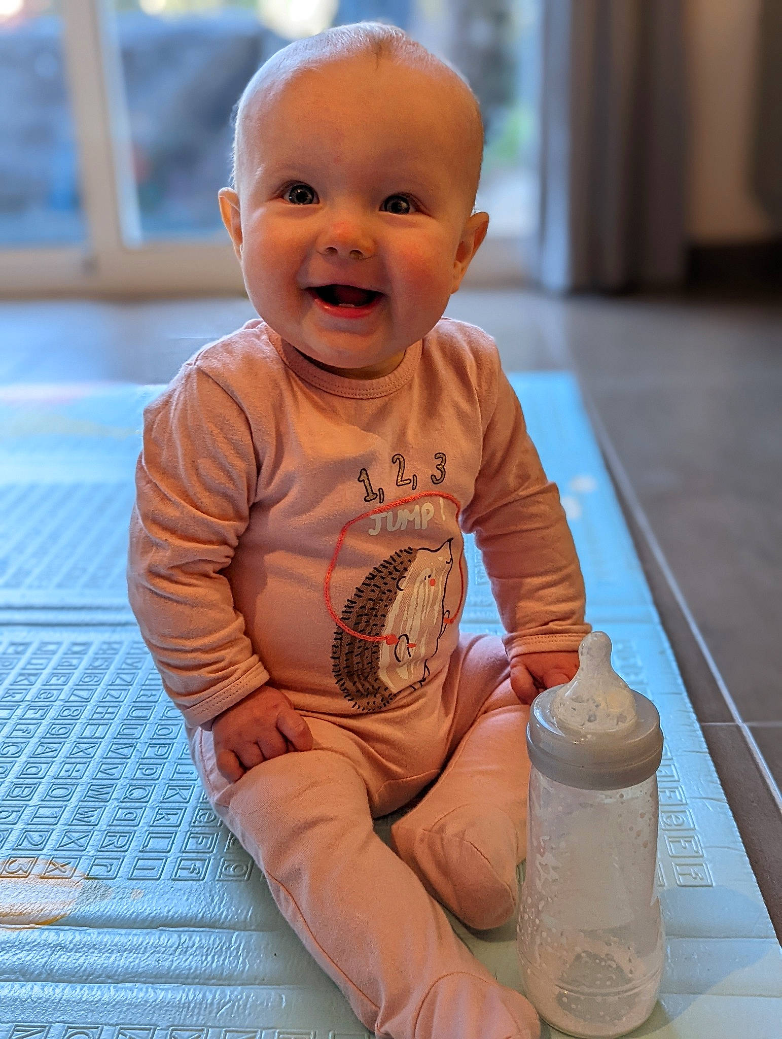 Naomi a rejoint le concours — aidez-le/la à gagner de superbes lots ! baby, baby_products, baby_toddler_clothing, cheek, child, face, facial_expression, floor, flooring, foot, happy, head, human_leg, person, plastic_bottle, sitting, skin, smile, toddler, water_bottle