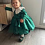 Branda Devert participe au concours pour gagner de l'argent avec cette photo : cabinet, child, curly_hair, cute, flooring, furniture, green_dress, green_shoes, hair_bow, home, indoor, person, playful, smiling, standing, toddler, white_collar, window, wood_floor, young_child