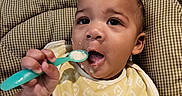 Legend is registered to the contest to win money with this photo: baby, bib, child, clothing, cute, dinnertime, expression, face, feeding, food, hand, high_chair, indoor, infant, kitchen, meal_time, seat, spoon, table, young_child