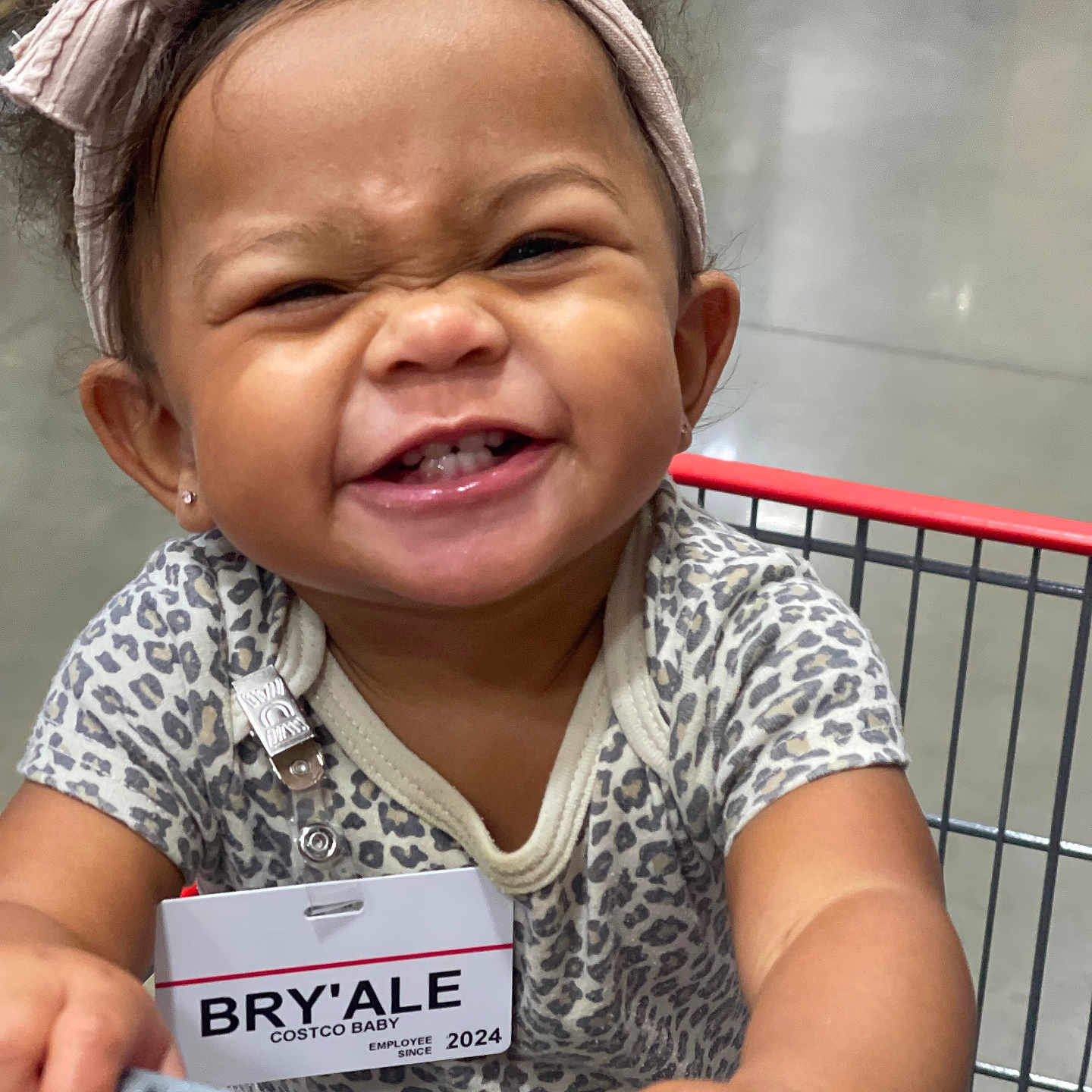 Bryale is registered to the contest to win money with this photo: accessory, baby, bow, child, clothing, cute, face, hands, happy, headband, holding, indoor, leopard_print, onesie, person, portrait, shopping, shopping_cart, smile, toddler