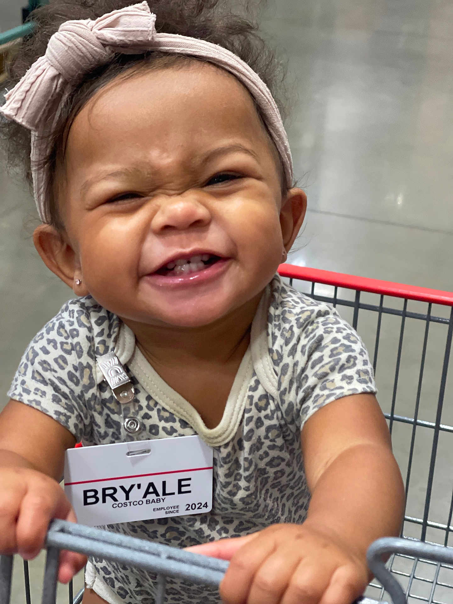 Bryale is registered to the contest to win money with this photo: accessory, baby, bow, child, clothing, cute, face, hands, happy, headband, holding, indoor, leopard_print, onesie, person, portrait, shopping, shopping_cart, smile, toddler