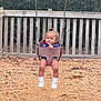 toddler, child, swing, playground, bucket_swing, wood_chips, wooden_fence, trees, outdoor, smiling, happy, casual_clothing, white_shoes, baby, fun, recreation, daytime, nature, park, person
