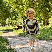 Maël participe au concours pour gagner de l'argent avec cette photo : child, toddler, curly_hair, striped_sweater, shorts, bottle, walking, path, park, greenery, trees, sunlight, daytime, outdoor, grass, nature, person, happy, cute, casual