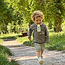 child, toddler, curly_hair, striped_sweater, shorts, bottle, walking, path, park, greenery, trees, sunlight, daytime, outdoor, grass, nature, person, happy, cute, casual