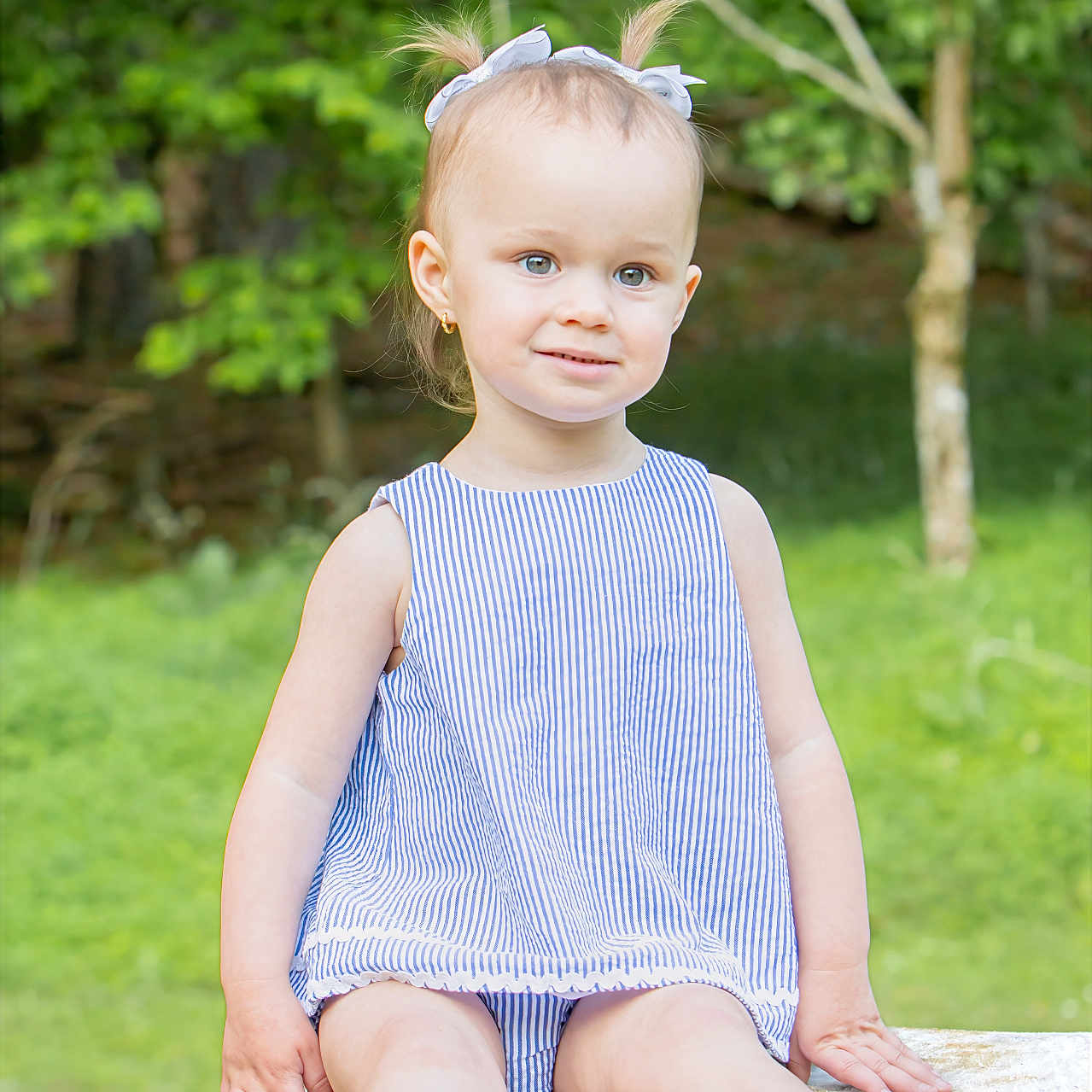 Everleigh joined the competition — help win amazing prizes! toddler, child, girl, outdoor, summer, greenery, tree_stump, dress, pigtails, smile, portrait, nature, grass, young_child, baby_sandals, cute, person, sitting, daylight, earrings