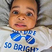 Adriel is registered to the contest to win money with this photo: baby, infant, child, face, eyes, tongue, clothing, shirt, text, graphic, sunglasses, blue, white, blanket, fabric, laying, portrait, cute, expression, indoors