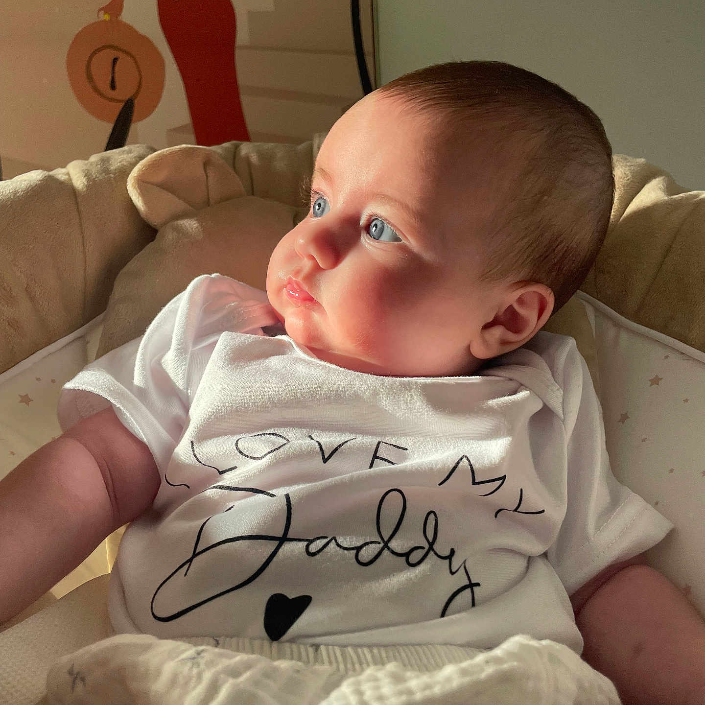 Alma a rejoint le concours — aidez-le/la à gagner de superbes lots ! baby, blue_eyes, child, closeup, cozy, cushion, cute, decor, expression, indoor, infant, person, portrait, relaxed, seated, side_profile, soft_lighting, text_on_clothing, wall_art, white_shirt