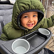 Saeed Campuzano is registered to the contest to win money with this photo: child, toddler, baby, smiling, happy, portrait, face, eyes, teeth, hood, jacket, winter_clothing, green, stroller, tray, cup_holder, outdoor, balcony, fence, close_up