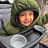child, toddler, baby, smiling, happy, portrait, face, eyes, teeth, hood, jacket, winter_clothing, green, stroller, tray, cup_holder, outdoor, balcony, fence, close_up
