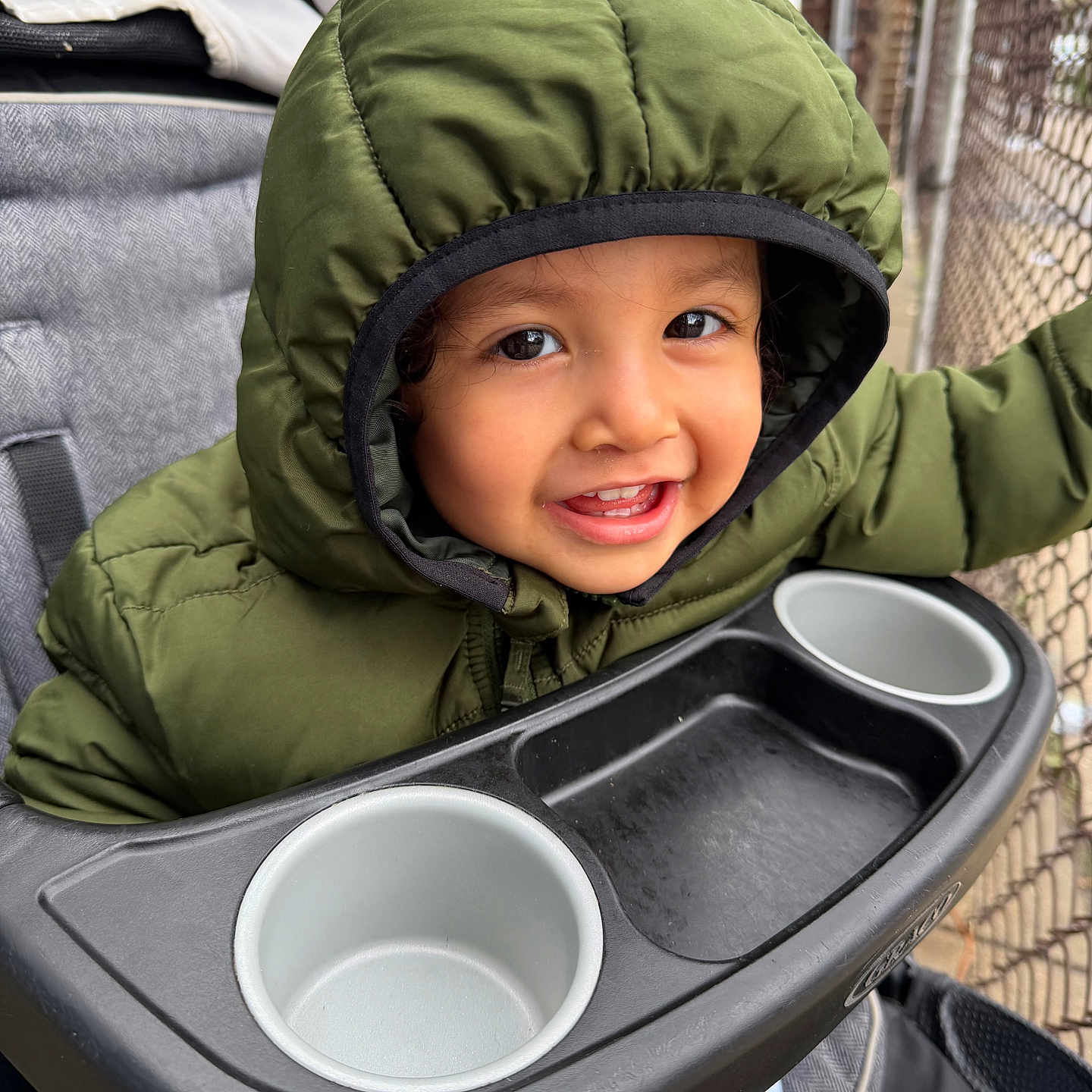 Saeed Campuzano is registered to the contest to win money with this photo: child, toddler, baby, smiling, happy, portrait, face, eyes, teeth, hood, jacket, winter_clothing, green, stroller, tray, cup_holder, outdoor, balcony, fence, close_up
