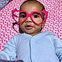 baby, infant, glasses, heart_shaped_glasses, pink, blue_clothing, blanket, patterned_blanket, cute, close_up, portrait, child, face, eyes, mouth, onesie, lying_down, indoor, soft_lighting, expression