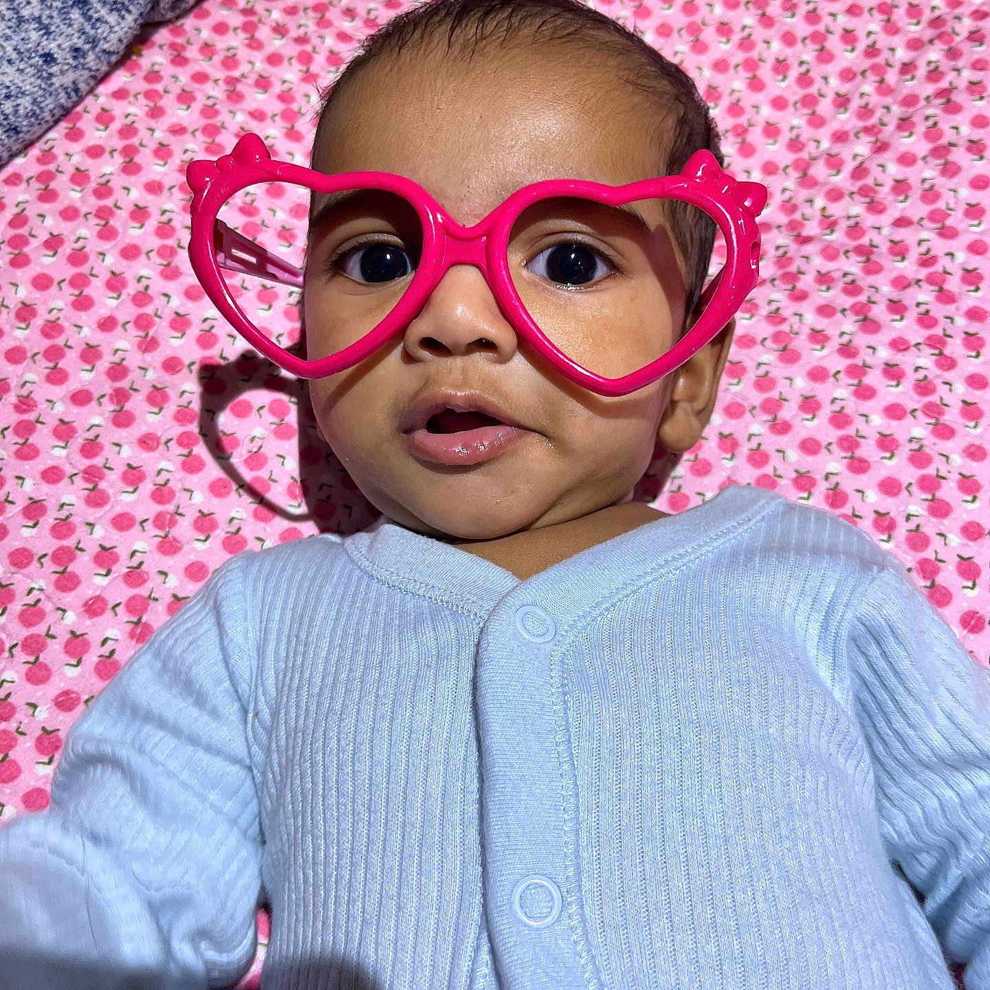 Adhyan is registered to the contest to win money with this photo: baby, blanket, blue_clothing, child, close_up, cute, expression, eyes, face, glasses, heart_shaped_glasses, indoor, infant, lying_down, mouth, onesie, patterned_blanket, pink, portrait, soft_lighting