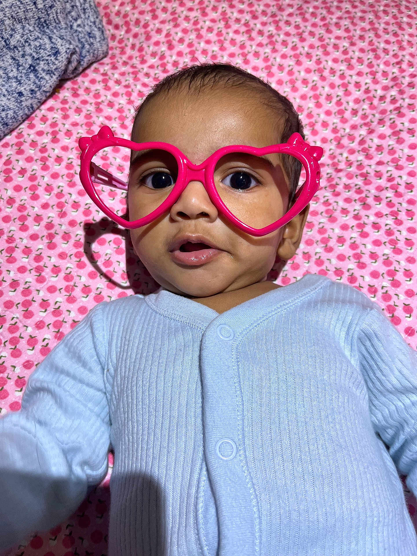 Adhyan is registered to the contest to win money with this photo: baby, infant, glasses, heart_shaped_glasses, pink, blue_clothing, blanket, patterned_blanket, cute, close_up, portrait, child, face, eyes, mouth, onesie, lying_down, indoor, soft_lighting, expression