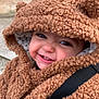 child, toddler, baby, smile, hood, fuzzy_coat, teddy_texture, closeup, portrait, rosy_cheeks, teeth, stroller_strap, warm_clothing, outdoor, stone_wall, candid, cute, eyes, playful_expression, coat