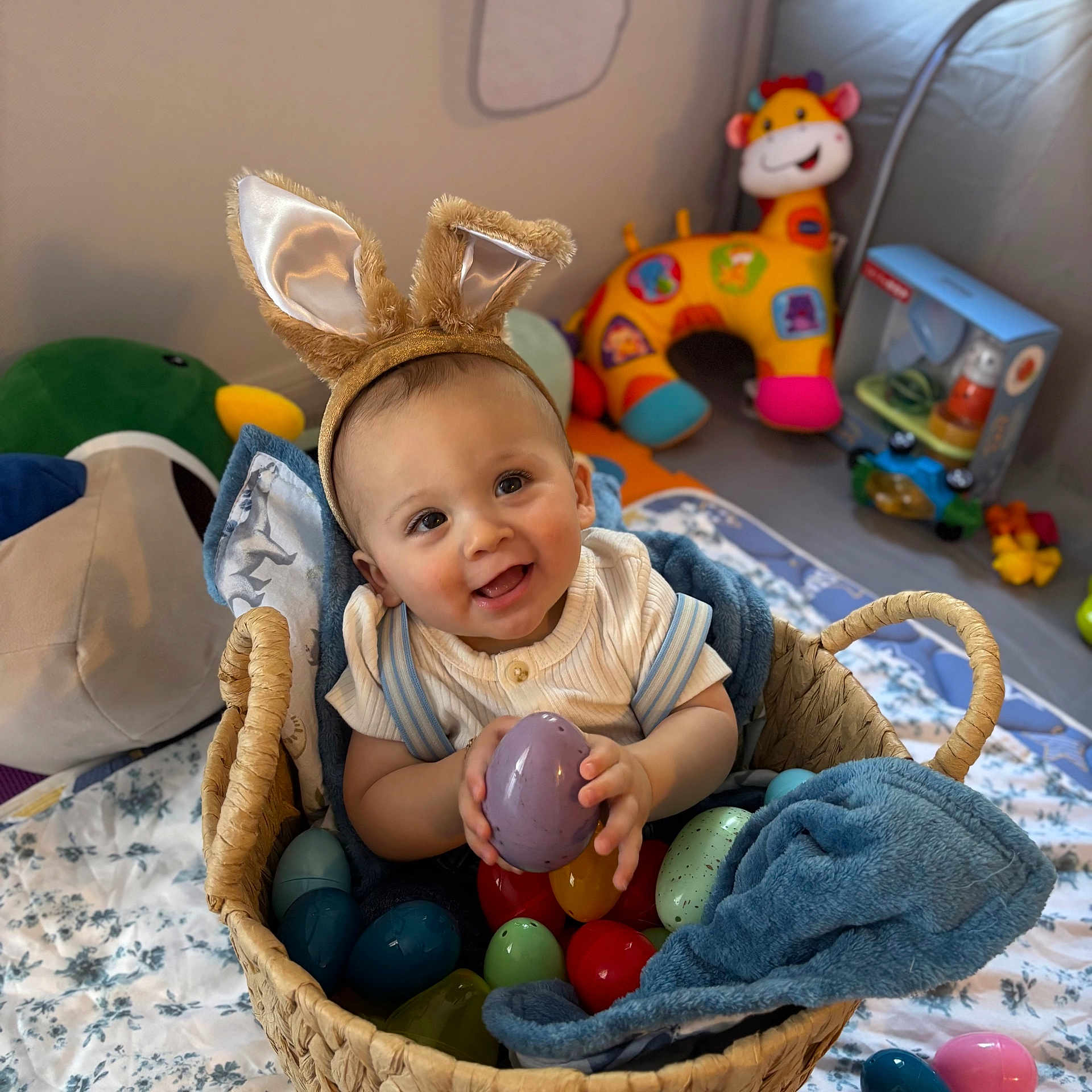 Audis is registered to the contest to win money with this photo: baby, bunny_ears, basket, plastic_eggs, blanket, plush_toys, toy_giraffe, toy_duck, smile, child, indoors, happy, cute, playroom, infant, fabric, headband, blue_blanket, woven_basket, colorful