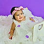 baby, child, infant, dress, white_dress, headband, pink_bow, purple_flowers, smiling, portrait, fluffy_surface, purple_background, cute, adorable, jewelry, bracelet, studio_photo, sitting, happy, young_child