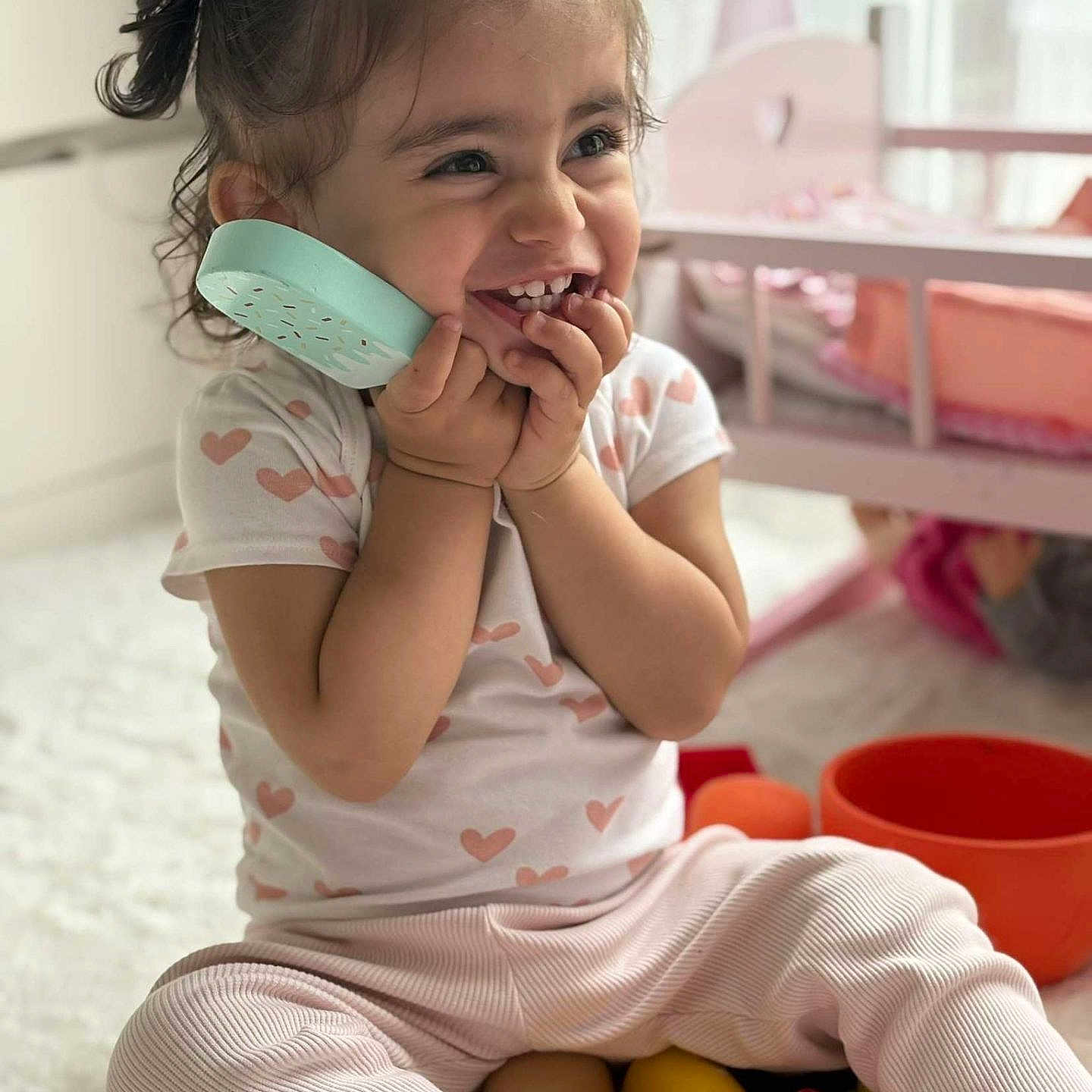 Aymila a rejoint le concours — aidez-le/la à gagner de superbes lots ! casual_clothing, child, cute, doll_crib, happy, heart_pattern, home, indoor, pigtails, pink_pants, playful, playtime, portrait, sitting, smiling, toddler, toy_phone, toys, white_rug, young_child