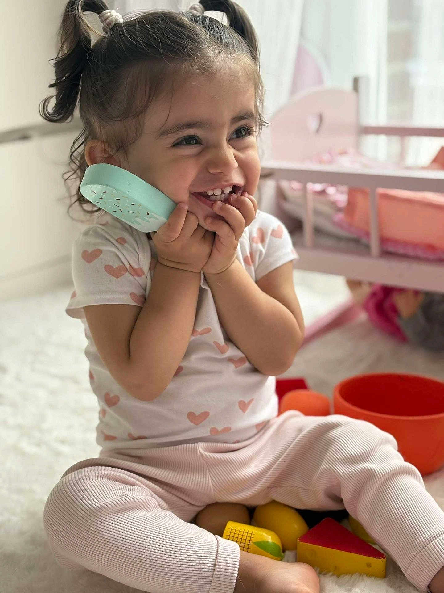 Aymila a rejoint le concours — aidez-le/la à gagner de superbes lots ! toddler, child, smiling, toy_phone, pigtails, heart_pattern, pink_pants, sitting, indoor, playtime, toys, doll_crib, white_rug, happy, cute, young_child, portrait, casual_clothing, home, playful