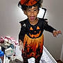 toddler, child, costume, halloween, pumpkin, hat, witch_hat, black_clothing, orange_clothing, smiling, jumping, indoor, changing_table, baby_care, happy, face, person, footwear, toy, blanket