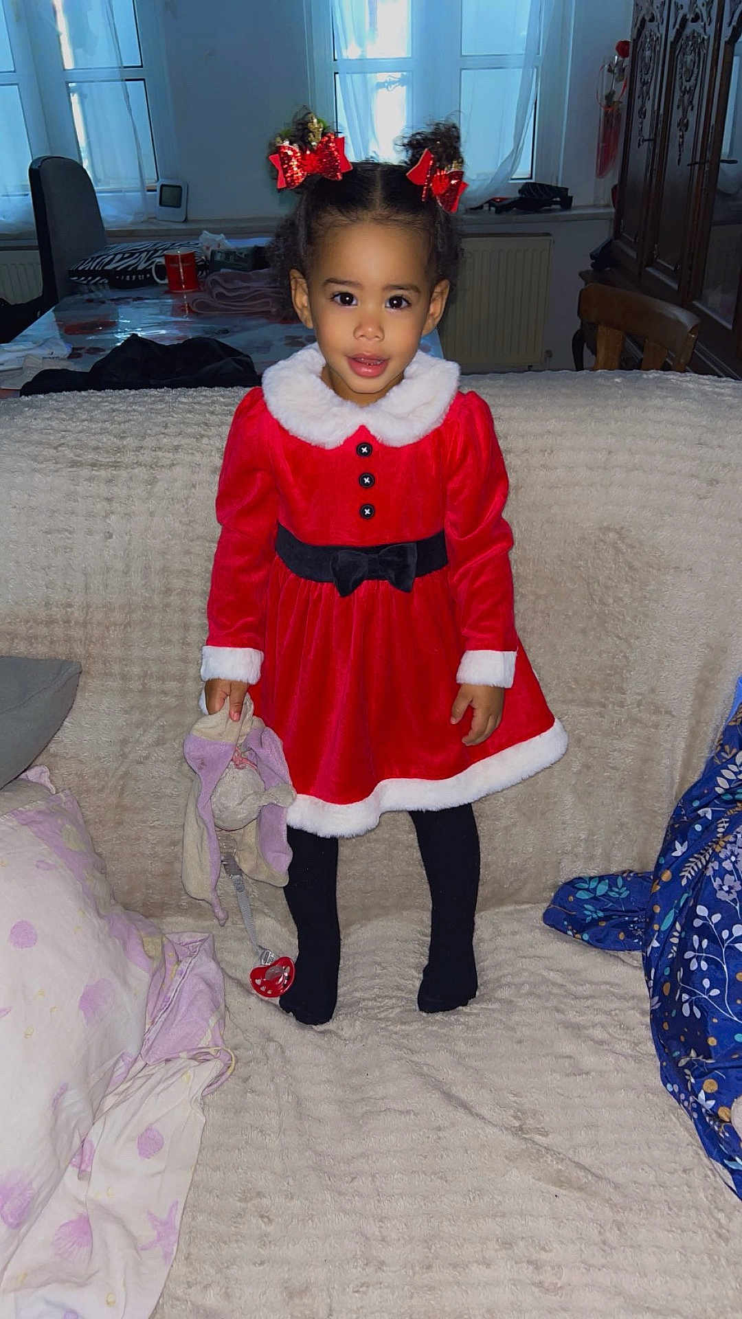 Maianna participe au concours pour gagner de l'argent avec cette photo : child, girl, red_dress, holiday_outfit, hair_bows, toy, pacifier, couch, blanket, indoor, smile, curly_hair, black_tights, cozy, home, furniture, window, curtains, chair, wooden_cabinet