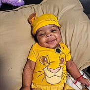Demetrius joined the competition — help win amazing prizes! baby, smiling, yellow_clothing, simba_outfit, hat, plush_toy, cushion, indoor, happy, child, portrait, cute, infant, person, lying_down, accessory, bracelet, toy, comfortable, relaxed