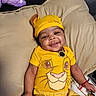 baby, smiling, yellow_clothing, simba_outfit, hat, plush_toy, cushion, indoor, happy, child, portrait, cute, infant, person, lying_down, accessory, bracelet, toy, comfortable, relaxed