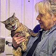 Charm is registered to the contest to win money with this photo: woman, cat, tabby_cat, door, indoor, person, holding, expression, feline, pet, animal, hand, clothing, jacket, face, teeth, mouth_open, fur, lock, handle