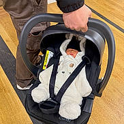 Skyler is registered to the contest to win money with this photo: adult, baby, bear_costume, black_hat, car_seat, child, clothing, floor, footwear, hand, indoor, infant, jacket, onesie, pants, person, safety, sleeping, straps, wooden_floor