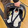 adult, baby, bear_costume, black_hat, car_seat, child, clothing, floor, footwear, hand, indoor, infant, jacket, onesie, pants, person, safety, sleeping, straps, wooden_floor