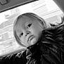 child, toddler, coat, blanket, car_interior, window, face, portrait, black_and_white, fur_collar, curious, young_child, clothing, seat, person, indoor, expression, soft_texture, automobile, travel