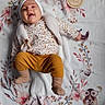 baby, infant, child, blanket, headband, clothing, lying_down, smiling, cute, soft_texture, wooden_sign, floral_pattern, mustard_pants, socks, portrait, indoors, adorable, happy, young_child, person