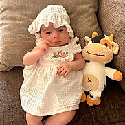 Aurora joined the competition — help win amazing prizes! baby, infant, white_dress, bonnet, plush_toy, cow_plush, couch, brown_couch, soft_fabric, cute, child, indoors, relaxed, portrait, smiling, toy, comfort, seated, looking_at_camera, cozy