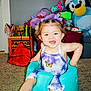 toddler, child, smiling, baby_seat, purple_bows, butterfly_outfit, toys, plush_toy, penguin, santa_hat, carpet, indoor, happy, cute, colorful, playroom, young_child, portrait, fun, seated