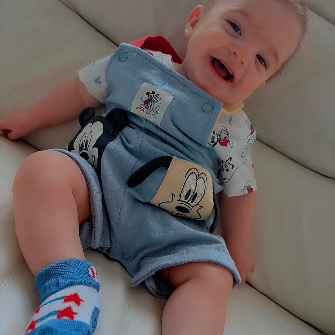 Carter is registered to the contest to win money with this photo: baby, child, smiling, mickey_mouse, overalls, socks, couch, indoor, happy, cute, infant, portrait, clothing, person, sitting, small_child, young, fun, playful, relaxed