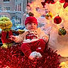 Carter is registered to the contest to win money with this photo: baby, christmas_tree, ornaments, red_hat, red_pants, plush_toy, grinch, window, cityscape, tinsel, festive_clothing, holiday_decor, lights, indoor, child, smiling, sitting, cozy, winter, cute
