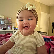 Katelyn joined the competition — help win amazing prizes! baby, bed, blanket, child, cozy, cute, face, flower, furniture, happy, headband, indoor, infant, person, portrait, room, sitting, smiling, striped_clothing, warm_lighting