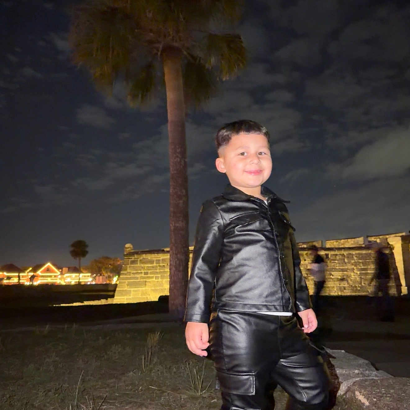Noham is registered to the contest to win money with this photo: boy, casual, child, clouds, confident, dark_sky, fashion, grass, leather_jacket, leather_pants, night, outdoor, palm_tree, person, portrait, smile, sneakers, stone_wall, urban, young