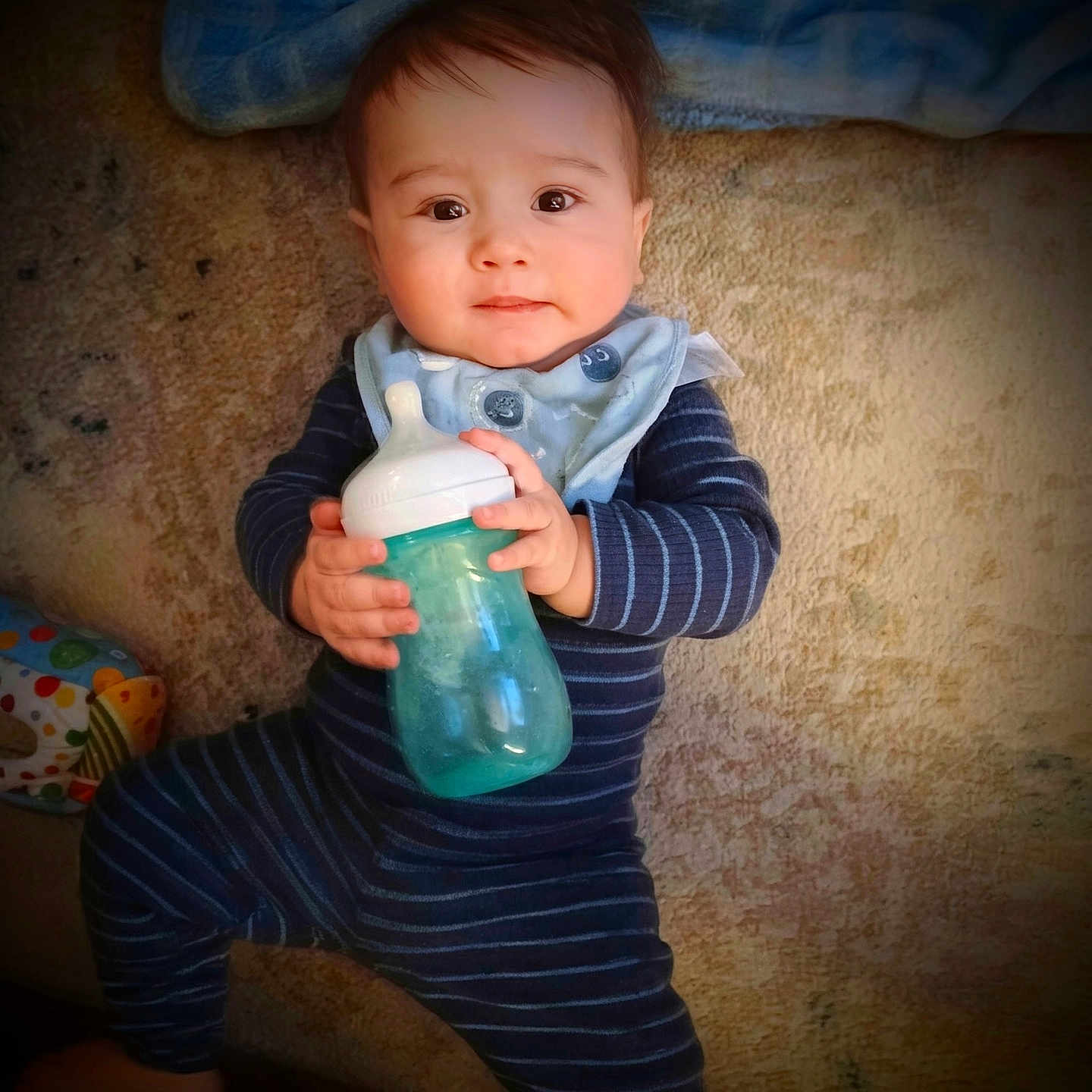 Gabriel joined the competition — help win amazing prizes! baby, blanket, blue, bottle, carpet, child, cute, face, floor, hand, home, indoor, infant, lying_down, pajamas, person, smile, soft_lighting, striped_clothing, teal