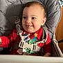 baby, infant, high_chair, safety_harness, buckle, holiday_sweater, christmas_tree, tongue_out, smile, face, eyes, hair, cheek, ear, plastic_tray, seat, indoor, wooden_background, portrait, cute