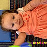 Isabel is registered to the contest to win money with this photo: child, toddler, girl, baby, portrait, face, eyes, looking_at_camera, dress, coral_clothing, embroidered_flowers, sitting, lying_down, classroom, carpet, blue_background, colorful, indoor, learning_environment, photograph