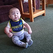 Lakoda is registered to the contest to win money with this photo: baby, child, bib, carpet, furniture, green_carpet, indoor, living_room, table, wooden_table, couch, plush_couch, clothing, socks, sitting, expression, person, face, cute, home