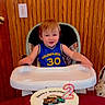 toddler, birthday_cake, high_chair, number_two_candle, golden_state_jersey, happy_birthday_balloon, wooden_wall, child, smiling, celebration, party, indoor, cake, table, red_tablecloth, person, face, clothing, birthday, food