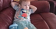 Lakoda is registered to the contest to win money with this photo: barefoot, casual, child, couch, cup, expression, face, furniture, indoor, lamp, leather, living_room, person, relaxing, shorts, sippy_cup, sitting, toddler, tshirt, wood_paneling